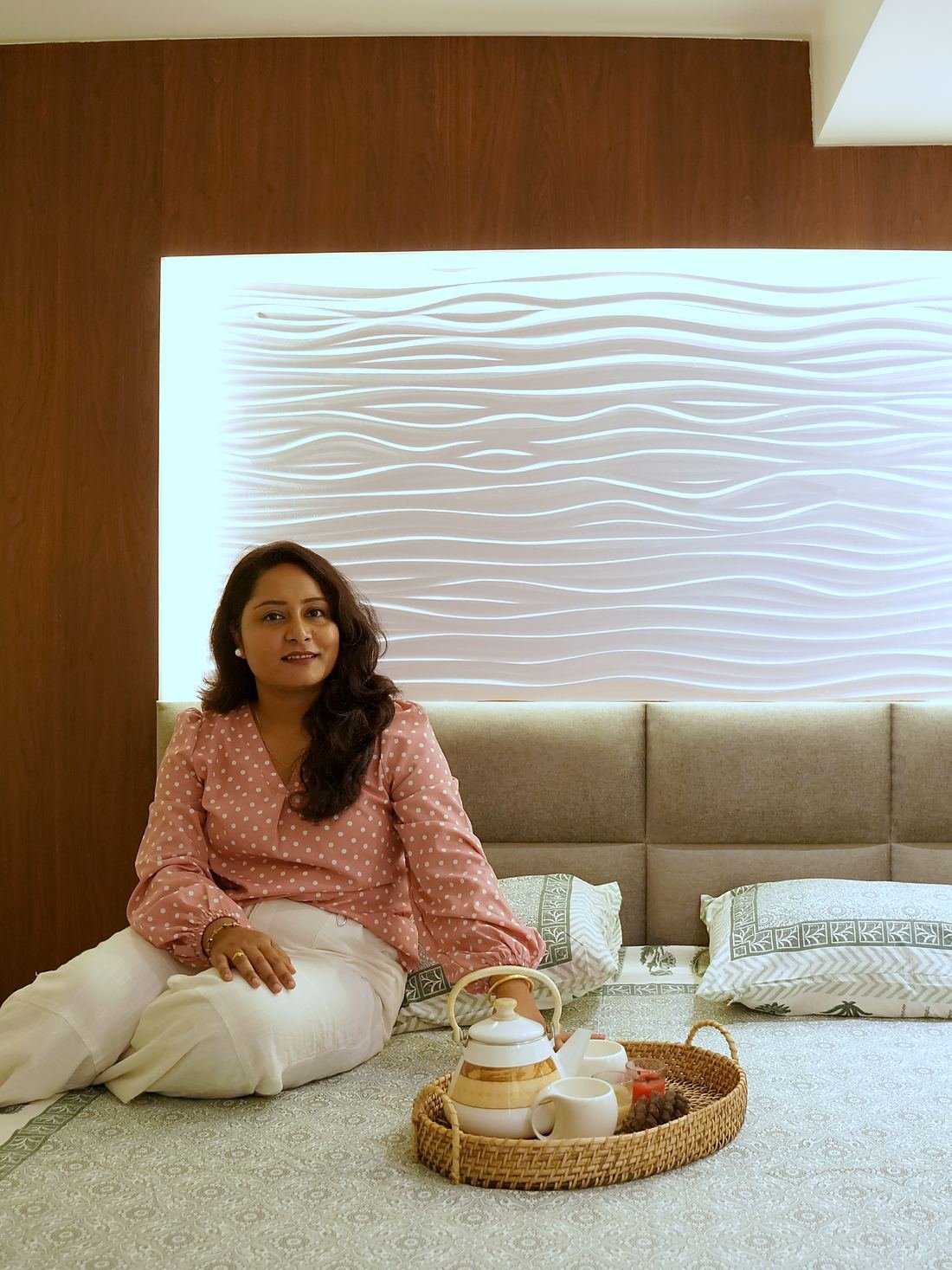 A guest pouring tea on the bed, framed by a backlit wave-textured headboard